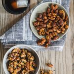 Two separate bowls hold two different recipes for Bourbon Spiced Nuts, one of which sits on a grey and white napkin while the other sits on the wood table.
