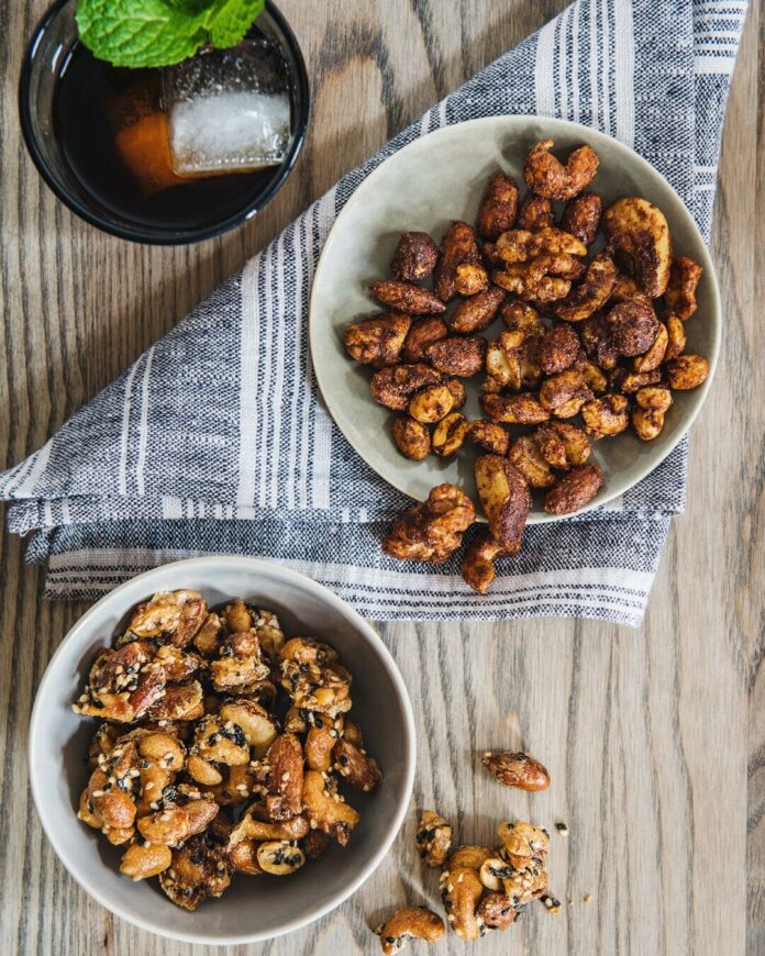 Two separate bowls hold two different recipes for Bourbon Spiced Nuts, one of which sits on a grey and white napkin while the other sits on the wood table.