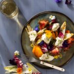A green olive plate holds a charred heirloom tomato and mozzarella salad with various bright vegetables and a glass of sparkling water nearby.