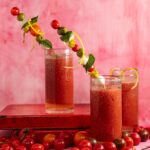 Three tall glasses filled with a vibrant red tomato-based drink, likely Bloody Marys, each garnished with skewers featuring colorful cherry tomatoes, mozzarella balls, green olives, basil leaves, and lemon zest spirals.