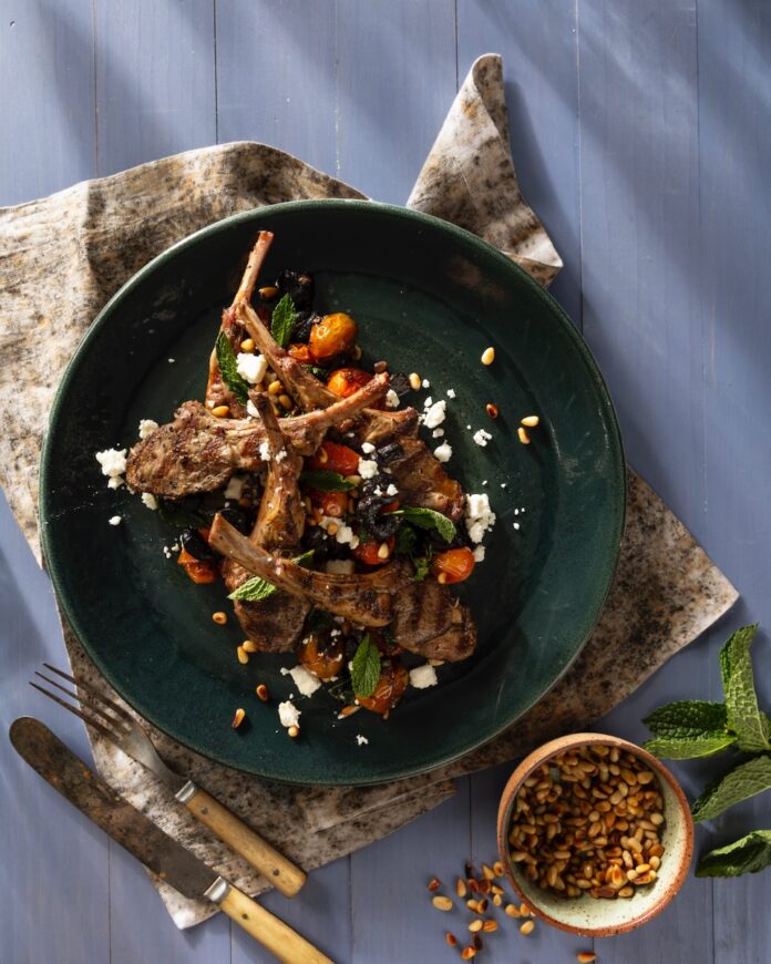 A green plate holds a grilled lamb legs scottadito with golden fork and knife beneath the plate and a small container of peppercorns on the other side.