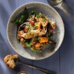 A white bowl full of a grilled Panzanella Salad with onions, tomatoes, and zucchini inside, all sitting on a blue picnic table with a spoon sitting below the bowl.