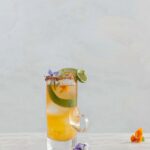 A Hot Clooney Mezcal Cocktail sits in a tall old fashioned glass with limes, tajin on the rim, and edible flowers on top.