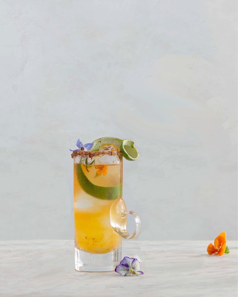 A Hot Clooney Mezcal Cocktail sits in a tall old fashioned glass with limes, tajin on the rim, and edible flowers on top.