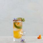 A Hot Clooney Mezcal Cocktail sits in a tall old fashioned glass with limes, tajin on the rim, and edible flowers on top.