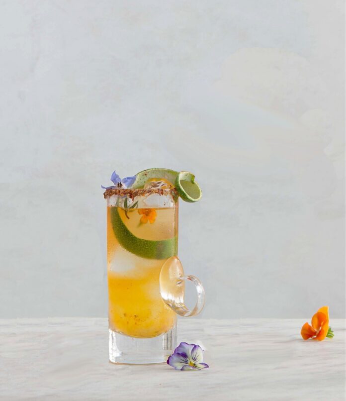 A Hot Clooney Mezcal Cocktail sits in a tall old fashioned glass with limes, tajin on the rim, and edible flowers on top.