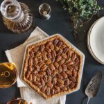 A hexagon shaped maple bourbon derby pie with pecans all over the top sits on a dark blue table with two cocktails to the left and white plates stacked to the right.