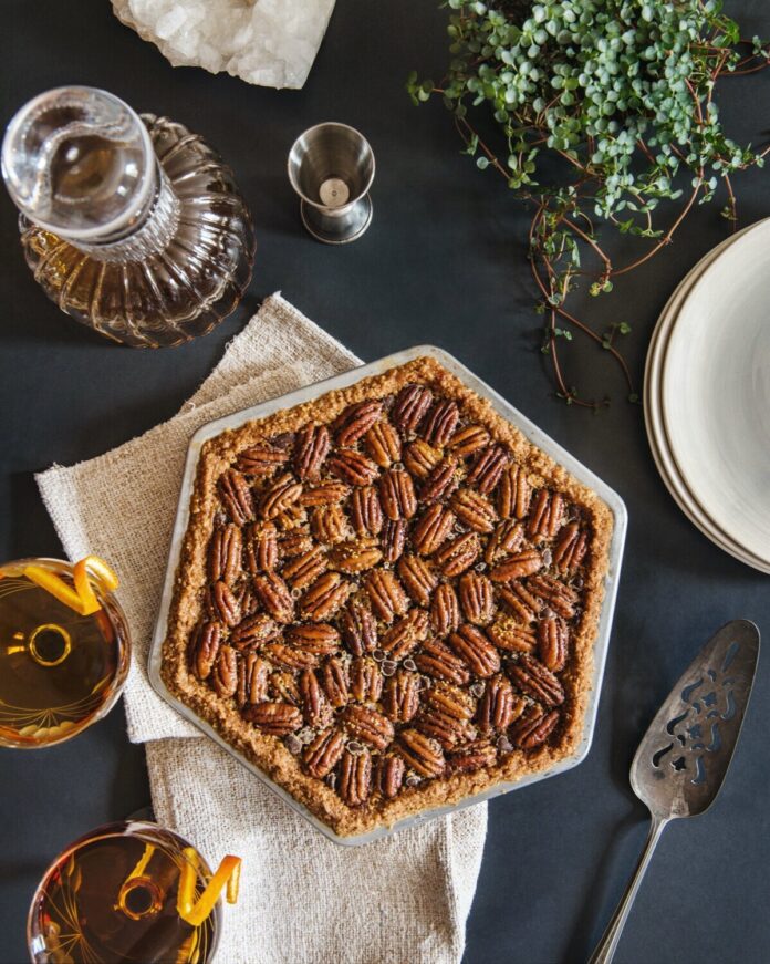 A hexagon shaped maple bourbon derby pie with pecans all over the top sits on a dark blue table with two cocktails to the left and white plates stacked to the right.