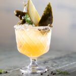 Lowball glass with a pickleback mocktail inside topped with pickle spears and kosher salt on the rim as it sits on a grey table.