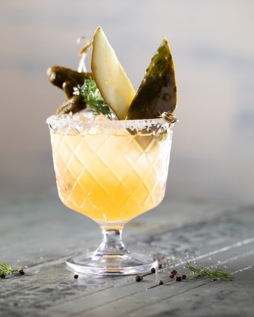 Lowball glass with a pickleback mocktail inside topped with pickle spears and kosher salt on the rim as it sits on a grey table.