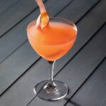 A cocktail glass with an orange rhubarb gimlet featuring a rhubarb ribbon garnish all on a black picnic table.