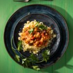 Two blue plates hold a tagine with chickpeas and rhubarb with a garnish of parsley underneath it, all on a green picnic table.