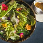 A salad with lettuce, radishes, edible flowers, and an apple bourbon dressing all in a black bowl on a dark blue table.