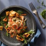 A dark plate holds garden vegetables and seafood in an escabeche salad with a fork and herbs to the right of the plate.