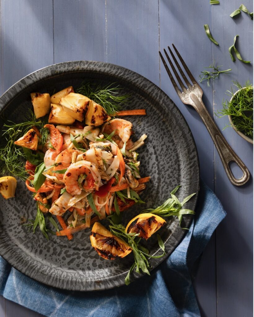 A dark plate holds garden vegetables and seafood in an escabeche salad with a fork and herbs to the right of the plate.