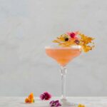 A tarragon gin and yellow chartreuse cocktail sits on a white marble table with edible flowers garnishing the glass and around the table.