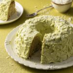 A round chiffon cake with coconut and match buttercream with a slice taken out of it that sits in the background.