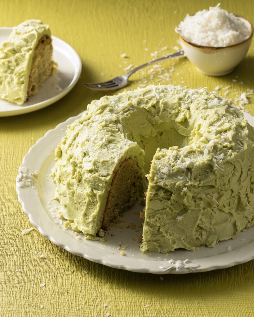 A round chiffon cake with coconut and match buttercream with a slice taken out of it that sits in the background.