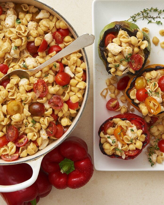 A white pot of Crab Pasta Salad with chunked tomatoes sits beside a white plate of grilled red pepper halves filled with the pasta salad.
