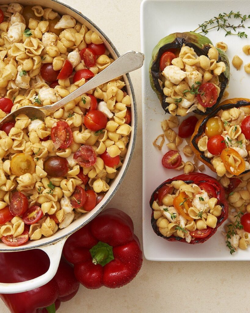 A white pot of Crab Pasta Salad with chunked tomatoes sits beside a white plate of grilled red pepper halves filled with the pasta salad.