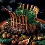 A crown roast of lamb with min sauce from the gilded age sits among a bed of figs and potatoes and greens.