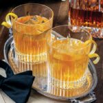 Two glasses of Fish House Punch with lemon twist garnishes sit on a silver serving tray.