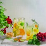 Two glasses of Fresh Summer Lemonade with oranges and limes sit in front of a pitcher of the lemonade as vines of green come in from the sides.