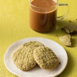 Three gluten-free matcha pistachio cookies on a plate on a green background with a cup of coffee above it.