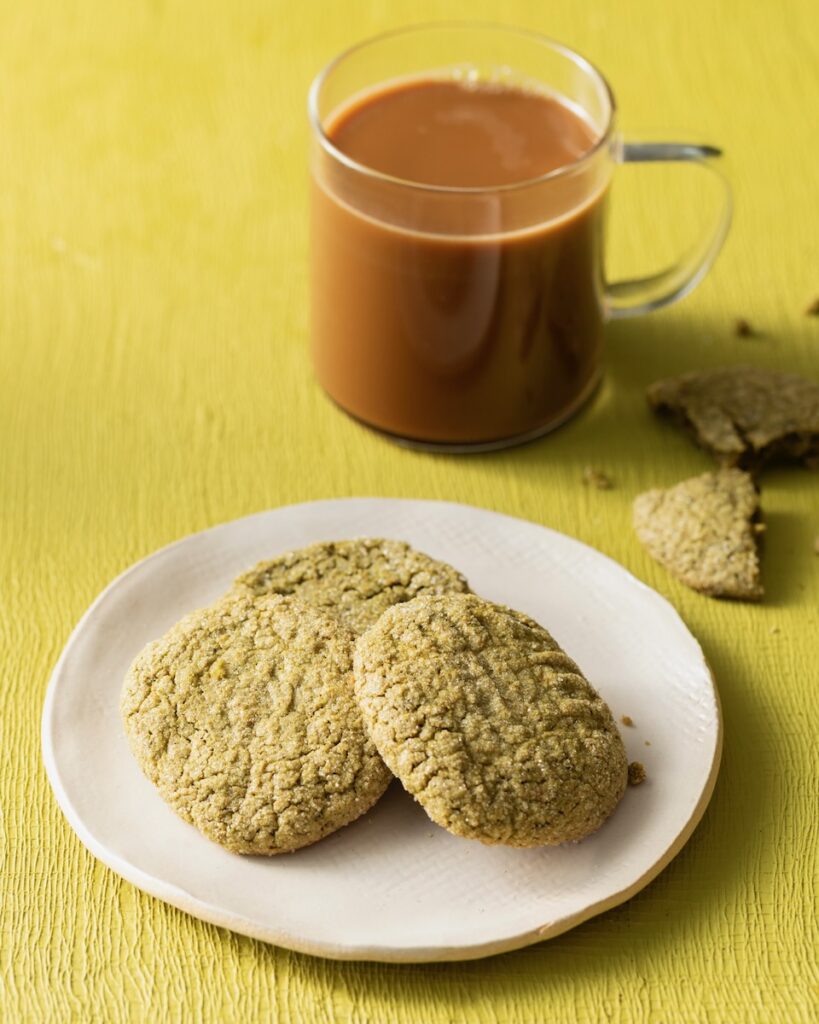 Three gluten-free matcha pistachio cookies on a plate on a green background with a cup of coffee above it.