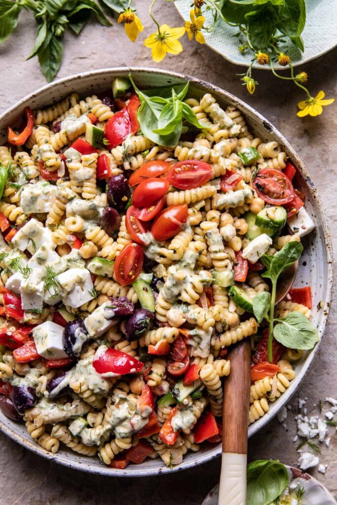 A Greek Pasta Salad sits in a grey bowl with pasta, tomatoes, cucumber, herbs, and feta cheese.