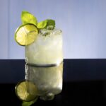 A green light cocktail with a lime circle and mint as garnishes against a black mirror surface.