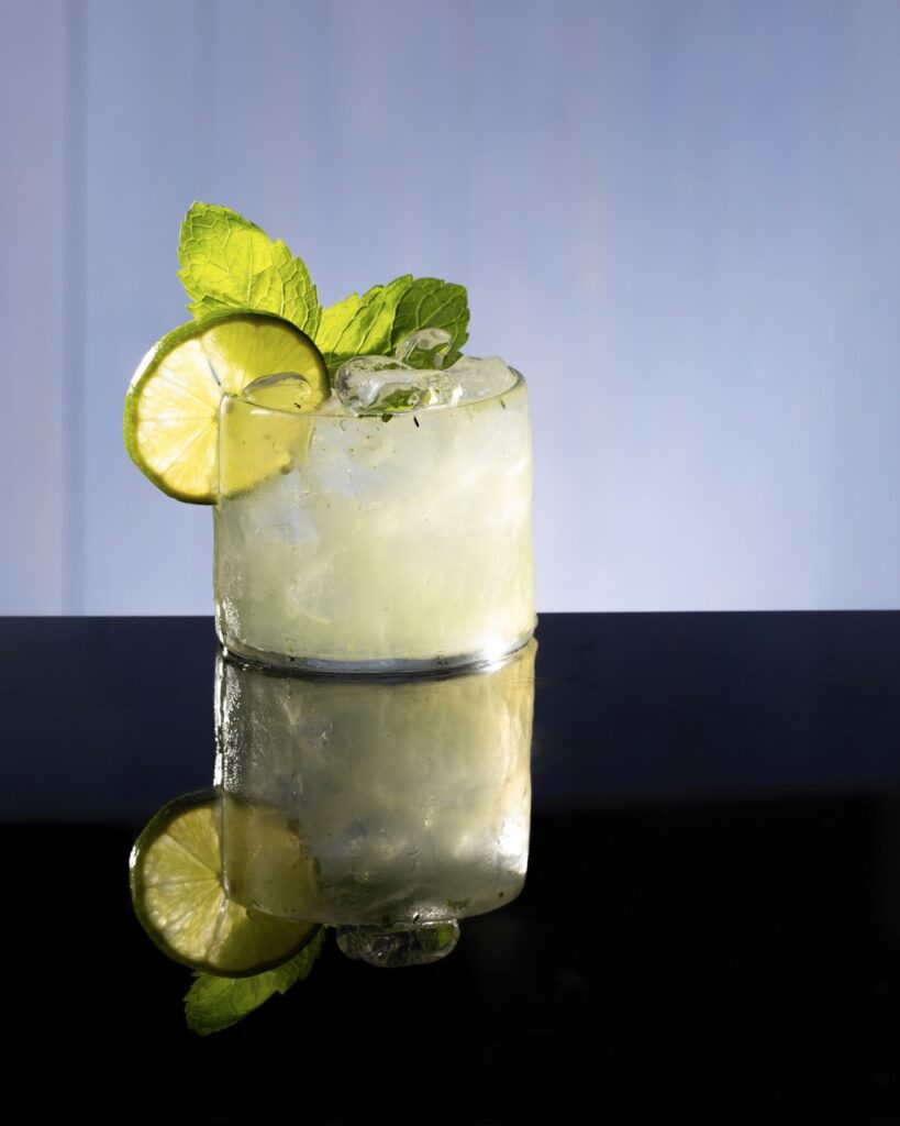 A green light cocktail with a lime circle and mint as garnishes against a black mirror surface.