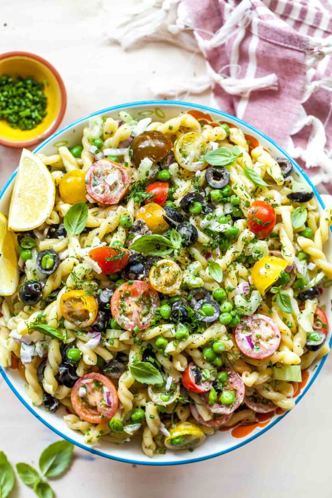 A blue bowl holds a Healthy Creamy Pasta Salad with tomatoes, fresh herbs, a slice of lemon and a small bowl of herbs in the upper left corner.