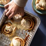 Four cinnamon rolls sit in a silver pan as a person reaching in to grab one. Two other cinnamon rolls sit on green plates in the top two corners.