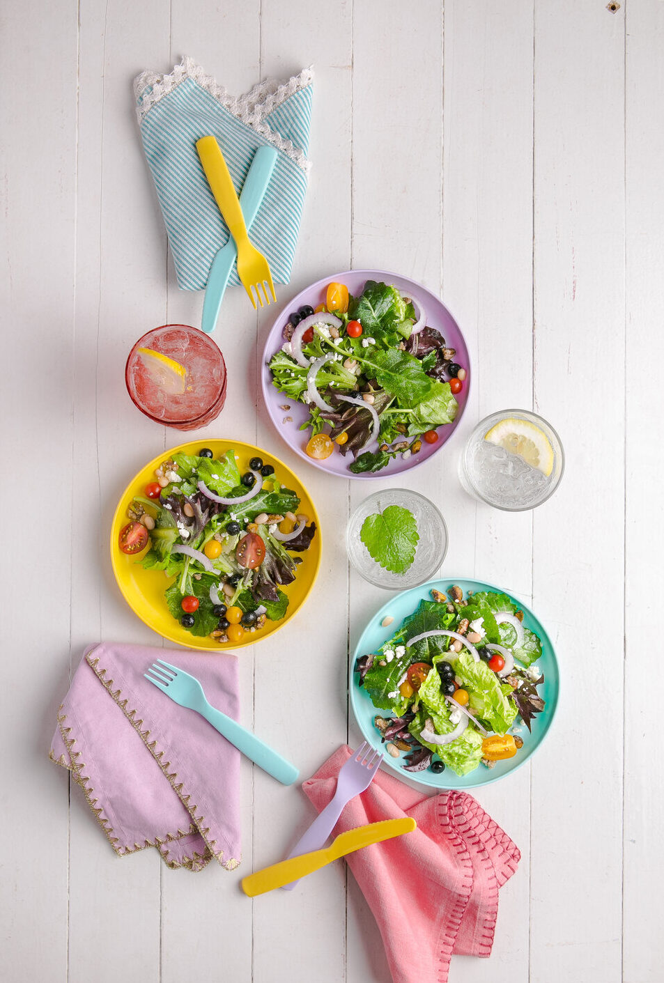 Three colorful bowls of kid-friendly summer salad with pink, purple, and blue napkins on the side.