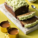 A loaf of matcha banana bread sits on a marble slab with the end in slices and a green buttercream on top. It all sits on a lime green background with two cups of coffee below it.