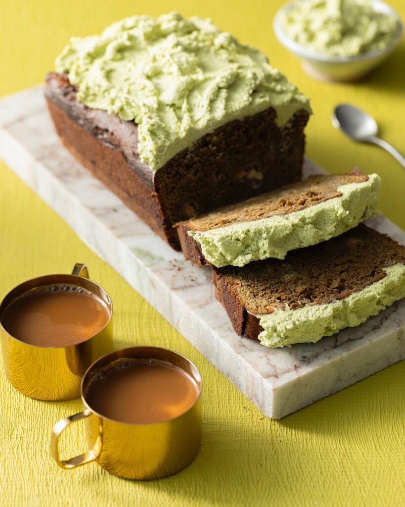 A loaf of matcha banana bread sits on a marble slab with the end in slices and a green buttercream on top. It all sits on a lime green background with two cups of coffee below it.