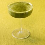 A coupe glass full of a green matcha martini with matcha powder on top all on the green background.