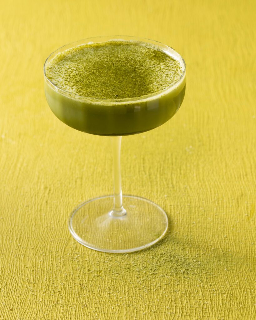 A coupe glass full of a green matcha martini with matcha powder on top all on the green background.