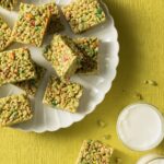 A white plate full of matcha rice krispie treats with mango. It sits beside two glasses of milk on a green background.