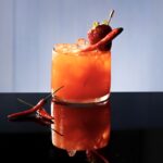A red light cocktail for squid game in orange color with chili peppers and a strawberry for garnish.