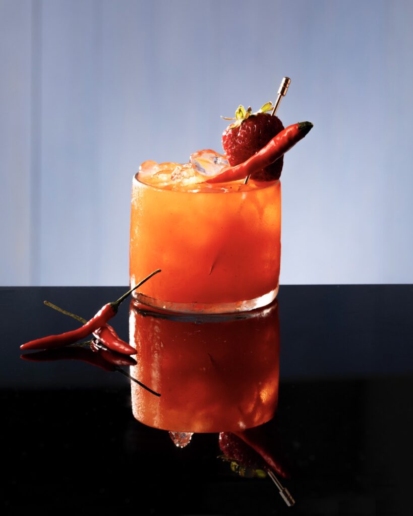 A red light cocktail for squid game in orange color with chili peppers and a strawberry for garnish.