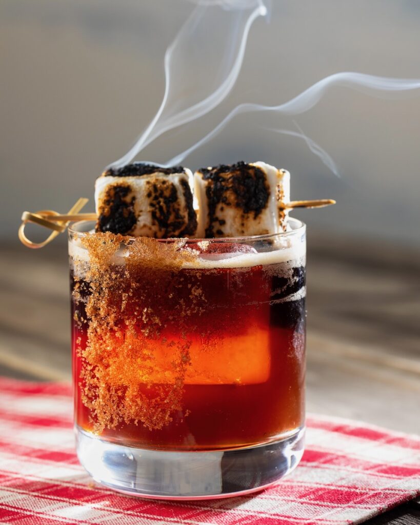 A short glass holds a S'mores Old Fashioned Mocktail featuring a skewer of toasted marshmallows on top and a rim of brown sugar.