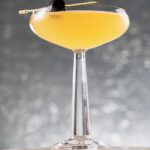 A yellow Superman's Kryptonite cocktail sits in a coupe glass with a toothpick with a black olive on it balancing on the edge. The glass sits on a stone block in front of a silver background.