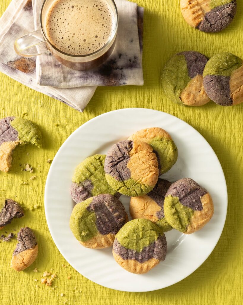 A white plate holds a stack of tricolor matcha sugar cookies in orange, purple, and green. The plate sits on a green background with a coffee above the plate and broken sugar cookies around it.