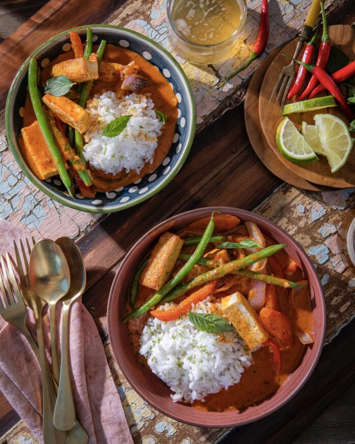 Two bowls of 20 minute coconut red curry sit on a table beside silverware, red chiles, lime slices, and a cup of beer.