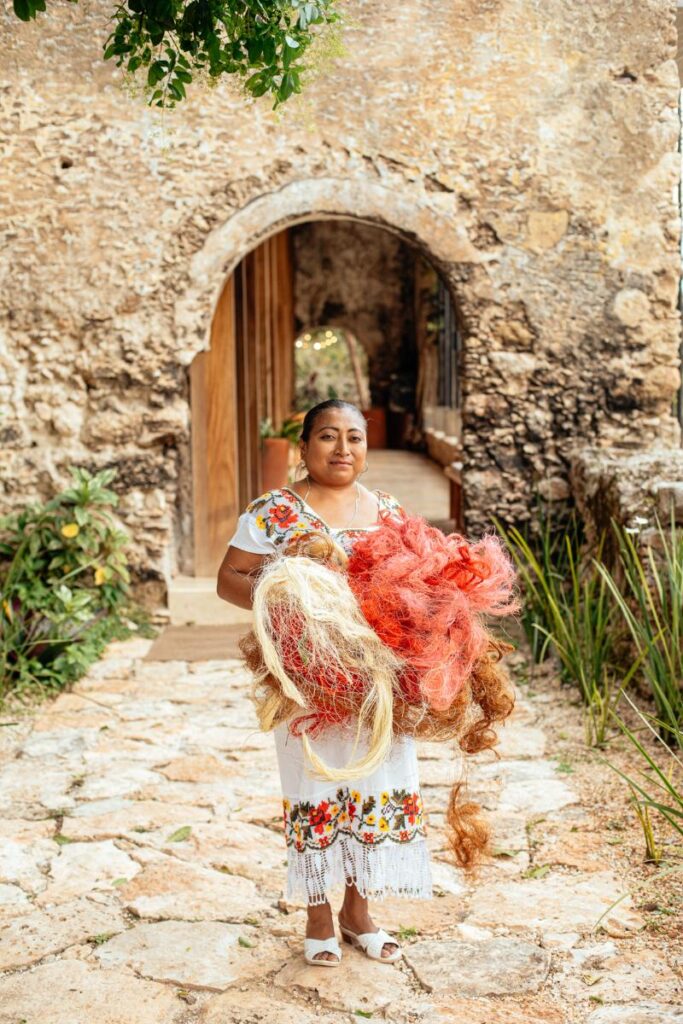 A woman wearing a traditional white dress with embroidered flowers holds a large bundle of red and light-colored fibers, standing in front of a stone archway.