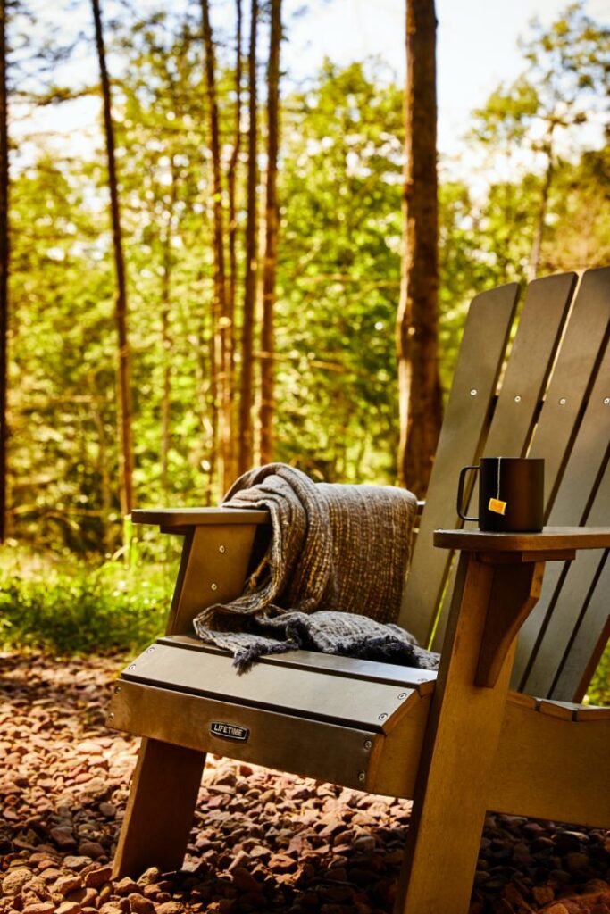 A dark green Adirondack chair with a brown and gray blanket draped over it sits on a rocky ground in a sunlit forest.