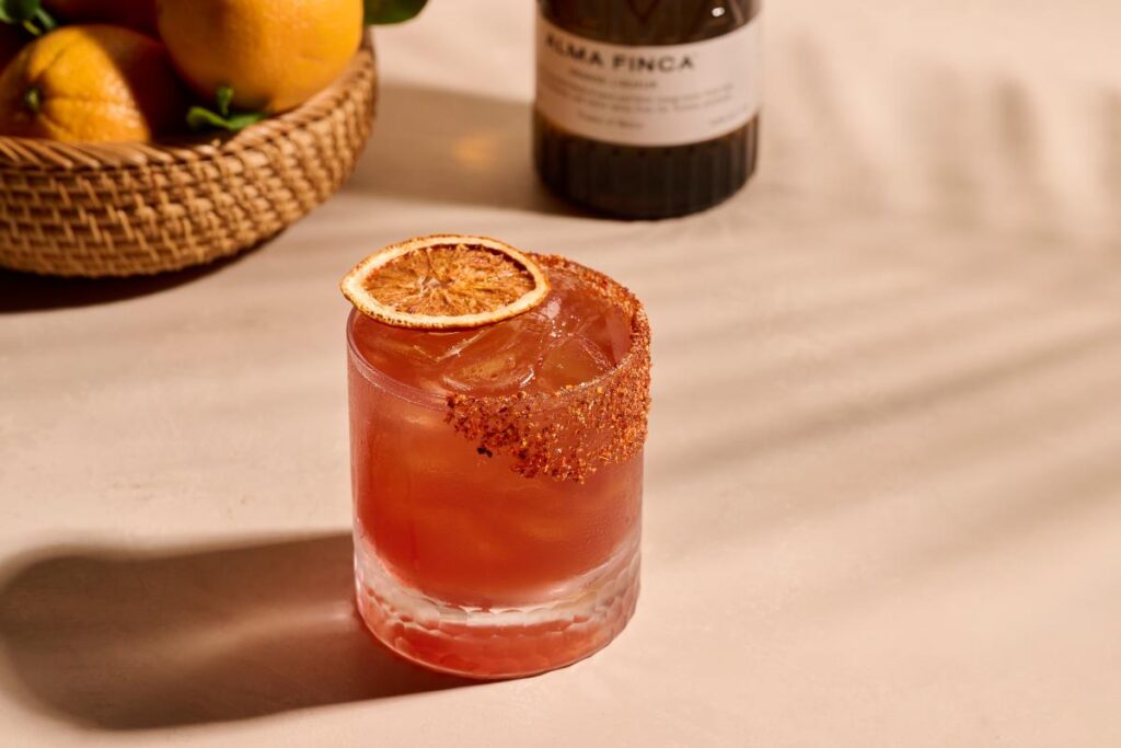 A clear glass filled with a pink-orange liquid and ice, garnished with a dried orange slice and a spiced rim.