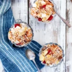 Three small cups of Ambrosia Salad with cherries on top sit on a white picnic table with a blue towel on it.